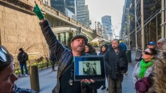 Justin Rivers, chief experience officer at Untapped New York, stands on a city sidewalk speaking to a group of participants while holding a tablet displaying a historic photograph. Tall buildings, scaffolding and pedestrians line the street in the background.