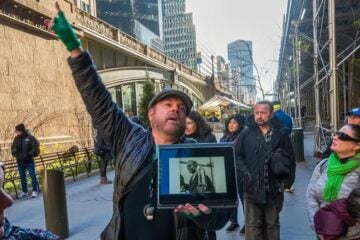 Justin Rivers, chief experience officer at Untapped New York, stands on a city sidewalk speaking to a group of participants while holding a tablet displaying a historic photograph. Tall buildings, scaffolding and pedestrians line the street in the background.