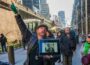 Justin Rivers, chief experience officer at Untapped New York, stands on a city sidewalk speaking to a group of participants while holding a tablet displaying a historic photograph. Tall buildings, scaffolding and pedestrians line the street in the background.