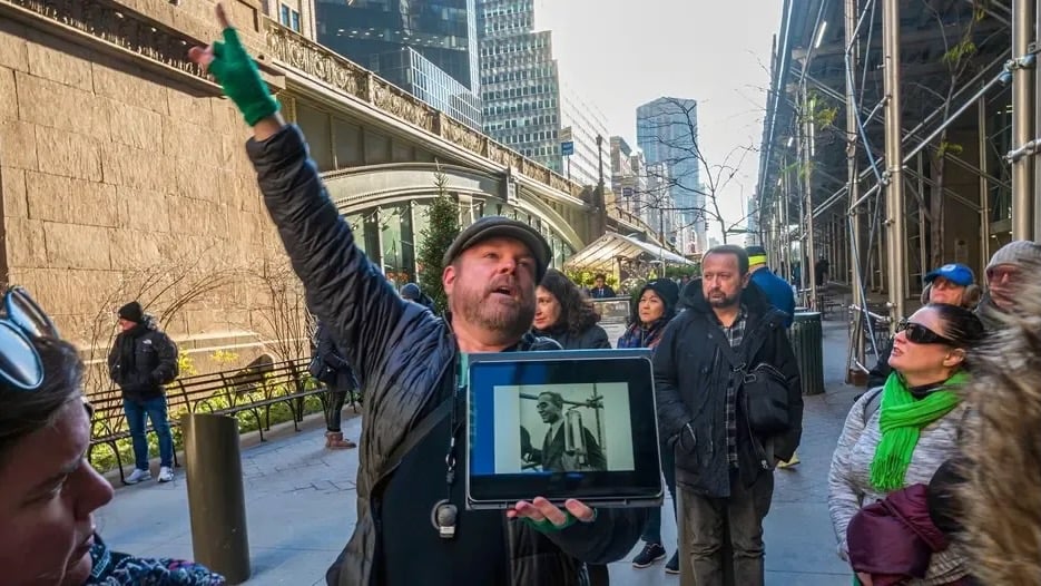 Justin Rivers, chief experience officer at Untapped New York, stands on a city sidewalk speaking to a group of participants while holding a tablet displaying a historic photograph. Tall buildings, scaffolding and pedestrians line the street in the background.