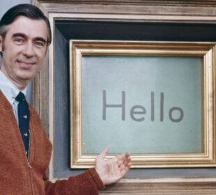 Mister Rogers stands beside his Picture Picture frame on the set of “Mister Rogers’ Neighborhood,” smiling and gesturing toward a screen displaying the word “Hello.” He wears a brown cardigan, white shirt and dark tie.