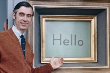 Mister Rogers stands beside his Picture Picture frame on the set of “Mister Rogers’ Neighborhood,” smiling and gesturing toward a screen displaying the word “Hello.” He wears a brown cardigan, white shirt and dark tie.