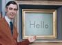 Mister Rogers stands beside his Picture Picture frame on the set of “Mister Rogers’ Neighborhood,” smiling and gesturing toward a screen displaying the word “Hello.” He wears a brown cardigan, white shirt and dark tie.