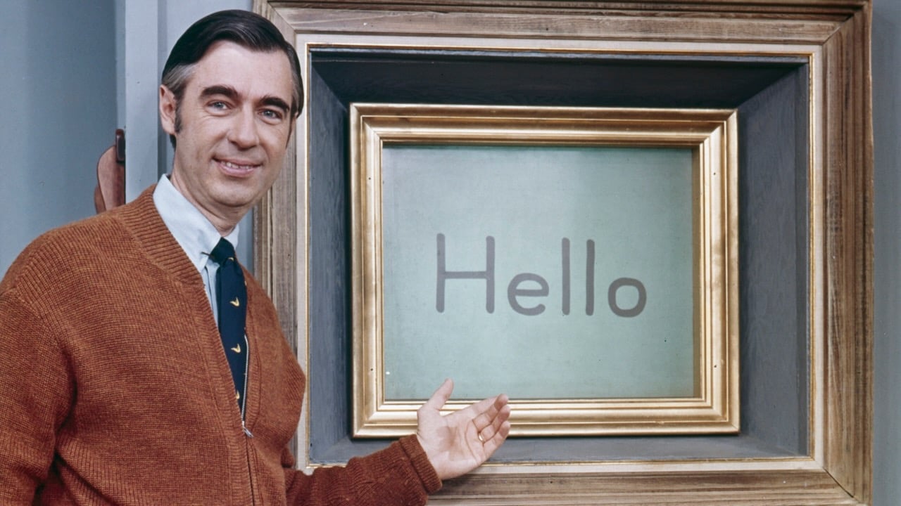 Mister Rogers stands beside his Picture Picture frame on the set of “Mister Rogers’ Neighborhood,” smiling and gesturing toward a screen displaying the word “Hello.” He wears a brown cardigan, white shirt and dark tie.
