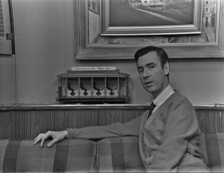 Black-and-white image of Fred Rogers seated on a plaid sofa on the set of the first episode of “Mister Rogers’ Neighborhood.” Behind him sits the show’s model trolley beneath a framed picture, as Rogers turns toward the camera wearing his trademark cardigan and tie.
