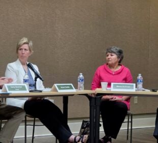Arkansas TV Commission Chairman Gary Newton, seated at left, speaks and gestures toward network CEO Carlton Wing, seated at right, during the commission’s June 4 meeting. Commissioners Anne Cowie and Maria Sullivan sit between them, listening as microphones, name placards and water bottles rest on the meeting tables.