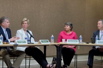 Arkansas TV Commission Chairman Gary Newton, seated at left, speaks and gestures toward network CEO Carlton Wing, seated at right, during the commission’s June 4 meeting. Commissioners Anne Cowie and Maria Sullivan sit between them, listening as microphones, name placards and water bottles rest on the meeting tables.