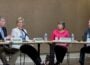 Arkansas TV Commission Chairman Gary Newton, seated at left, speaks and gestures toward network CEO Carlton Wing, seated at right, during the commission’s June 4 meeting. Commissioners Anne Cowie and Maria Sullivan sit between them, listening as microphones, name placards and water bottles rest on the meeting tables.