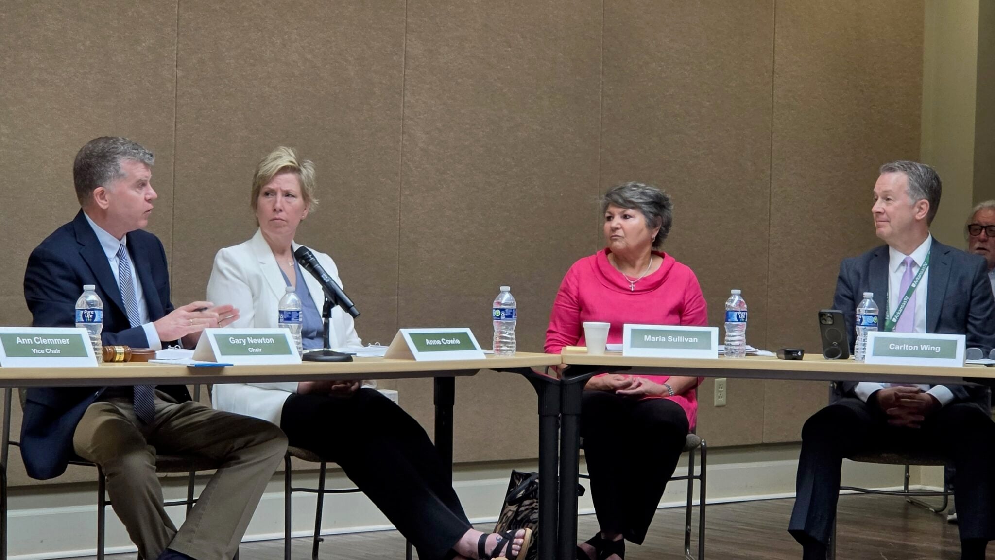 Arkansas TV Commission Chairman Gary Newton, seated at left, speaks and gestures toward network CEO Carlton Wing, seated at right, during the commission’s June 4 meeting. Commissioners Anne Cowie and Maria Sullivan sit between them, listening as microphones, name placards and water bottles rest on the meeting tables.