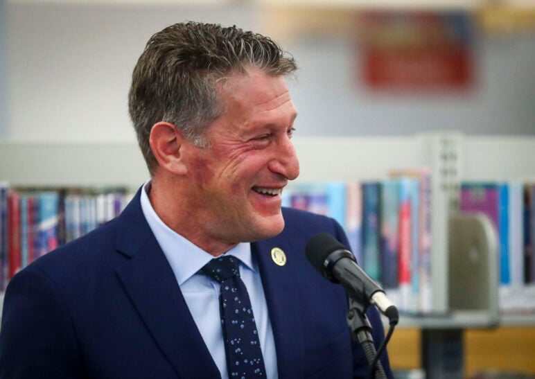 New Jersey state Sen. Andrew Zwicker smiles while speaking into a microphone in front of blurred bookshelves.