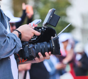 A filmmaker holds a professional video camera equipped with a microphone, monitor and matte box while recording at an outdoor event. Several people and camera tripods are visible out of focus in the background.