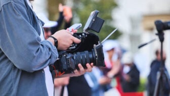 A filmmaker holds a professional video camera equipped with a microphone, monitor and matte box while recording at an outdoor event. Several people and camera tripods are visible out of focus in the background.