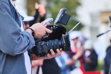 A filmmaker holds a professional video camera equipped with a microphone, monitor and matte box while recording at an outdoor event. Several people and camera tripods are visible out of focus in the background.