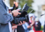 A filmmaker holds a professional video camera equipped with a microphone, monitor and matte box while recording at an outdoor event. Several people and camera tripods are visible out of focus in the background.