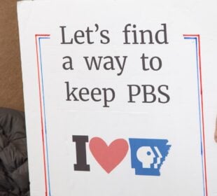 A person holds a handmade sign that reads “Let’s find a way to keep PBS” above an “I heart Arkansas PBS” graphic. Only the person’s torso and hand are visible. The sign is outlined in red and blue.