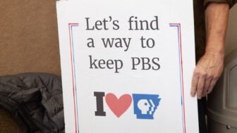 A person holds a handmade sign that reads “Let’s find a way to keep PBS” above an “I heart Arkansas PBS” graphic. Only the person’s torso and hand are visible. The sign is outlined in red and blue.