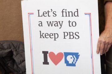 A person holds a handmade sign that reads “Let’s find a way to keep PBS” above an “I heart Arkansas PBS” graphic. Only the person’s torso and hand are visible. The sign is outlined in red and blue.