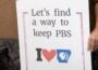 A person holds a handmade sign that reads “Let’s find a way to keep PBS” above an “I heart Arkansas PBS” graphic. Only the person’s torso and hand are visible. The sign is outlined in red and blue.