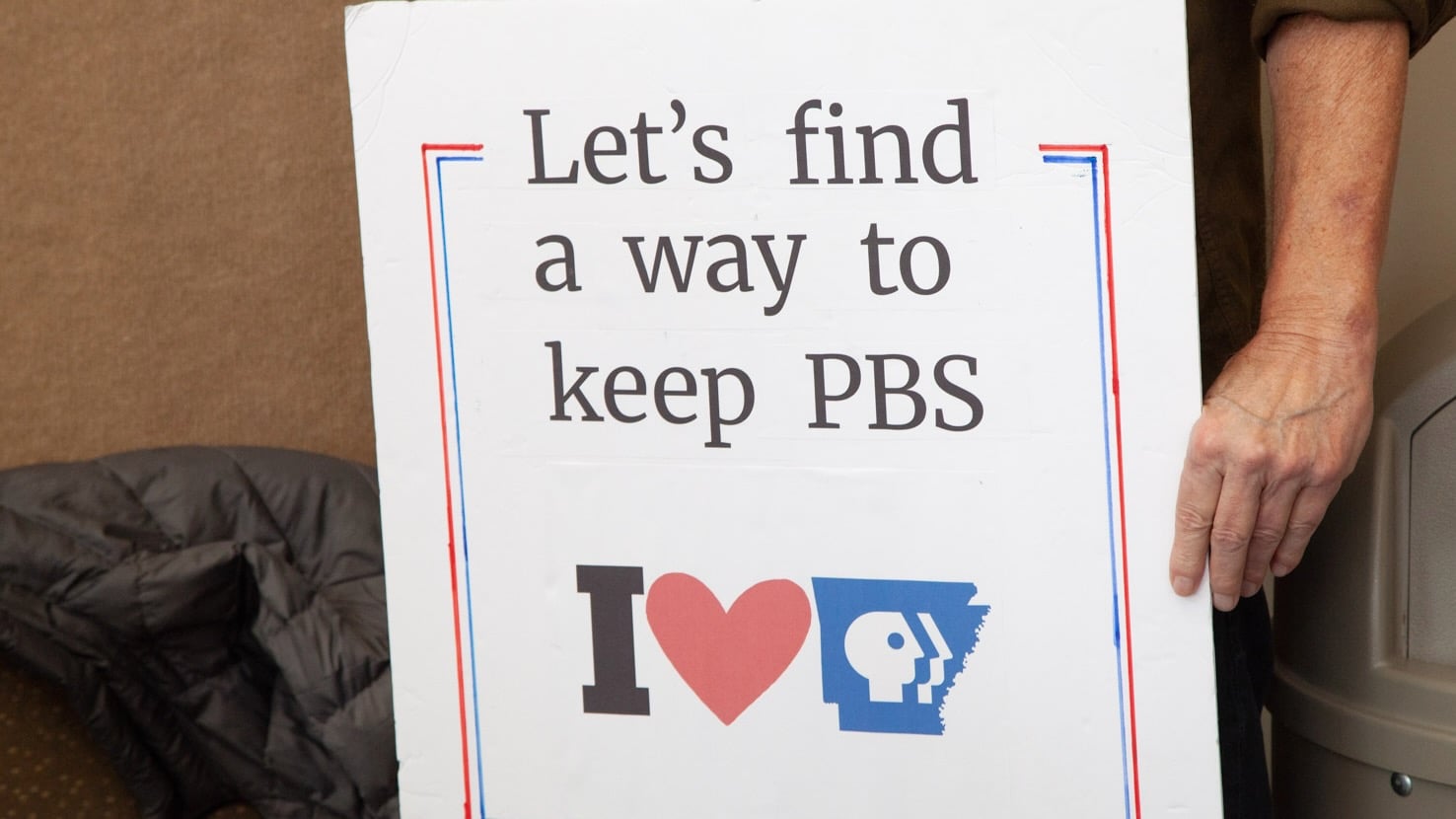A person holds a handmade sign that reads “Let’s find a way to keep PBS” above an “I heart Arkansas PBS” graphic. Only the person’s torso and hand are visible. The sign is outlined in red and blue.