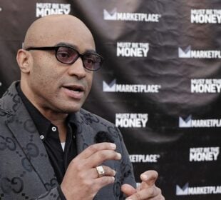 Lee Hawkins, host of the podcast Must Be the Money, speaks into a handheld microphone during an interview outside the podcast’s launch event at the Miracle Theater in Inglewood, California. He wears a patterned gray blazer and dark glasses, with a backdrop displaying Marketplace and Must Be the Money logos.