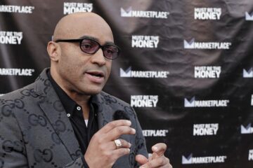 Lee Hawkins, host of the podcast Must Be the Money, speaks into a handheld microphone during an interview outside the podcast’s launch event at the Miracle Theater in Inglewood, California. He wears a patterned gray blazer and dark glasses, with a backdrop displaying Marketplace and Must Be the Money logos.