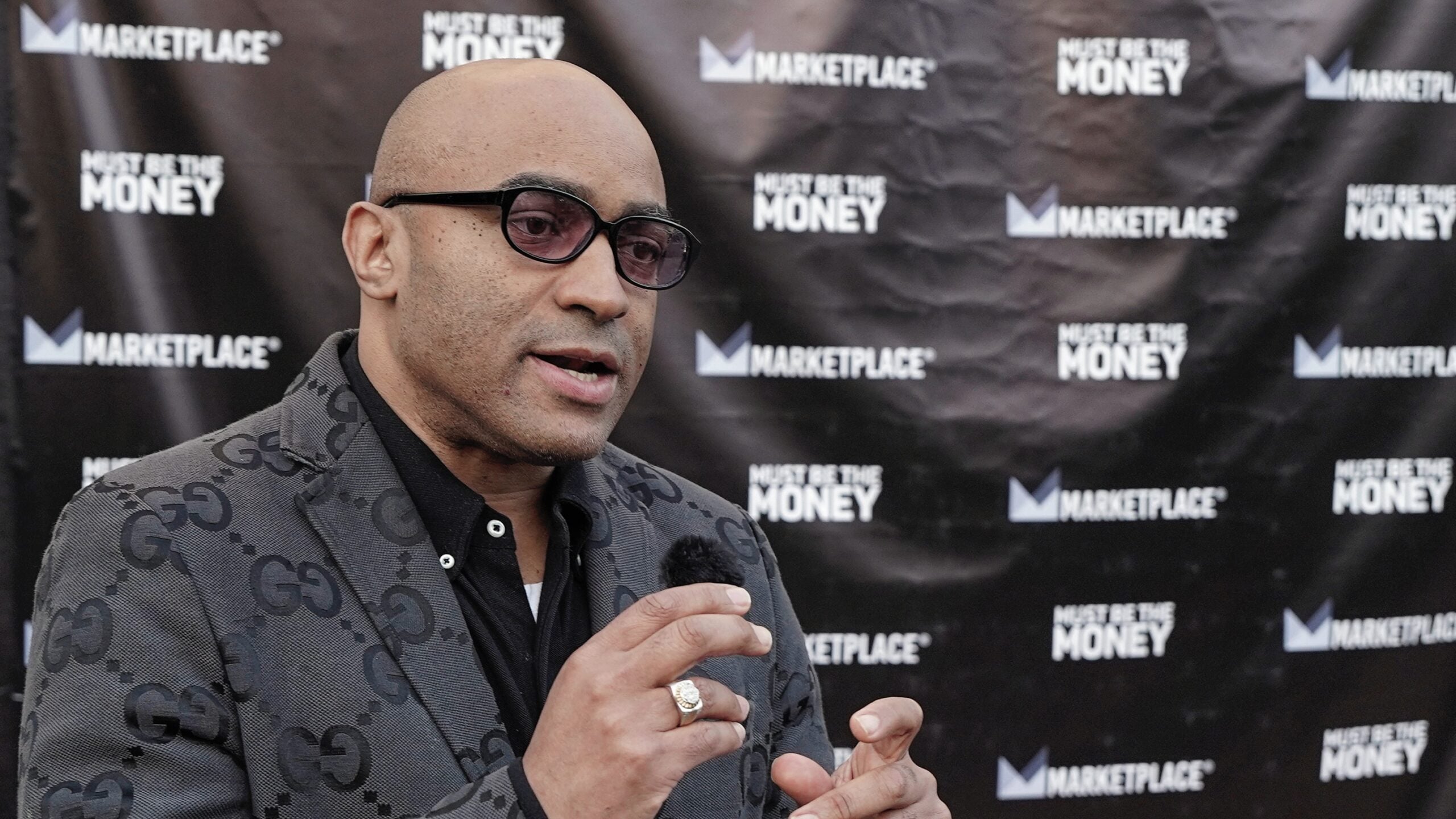 Lee Hawkins, host of the podcast Must Be the Money, speaks into a handheld microphone during an interview outside the podcast’s launch event at the Miracle Theater in Inglewood, California. He wears a patterned gray blazer and dark glasses, with a backdrop displaying Marketplace and Must Be the Money logos.