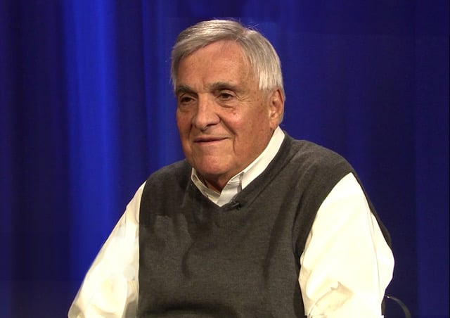 Mike LaBonia sits in a television studio wearing a button-down shirt and a gray sweater vest. He faces slightly to the side with a faint smile against a blue backdrop.