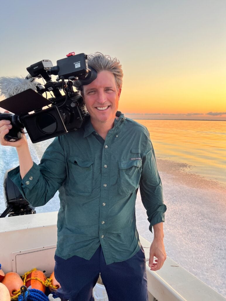 Patrick Greene, founder of Symbio Studios, stands on a boat at sunset, smiling while holding a professional video camera on his shoulder. Calm water and a colorful orange-and-gold sky stretch behind him.