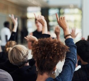 A diverse audience sits facing a speaker at a community event, with several attendees raising their hands to ask questions or participate in the discussion. The speaker is intentionally out of focus, emphasizing audience participation and engagement.