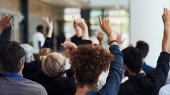 A diverse audience sits facing a speaker at a community event, with several attendees raising their hands to ask questions or participate in the discussion. The speaker is intentionally out of focus, emphasizing audience participation and engagement.