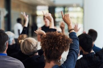 A diverse audience sits facing a speaker at a community event, with several attendees raising their hands to ask questions or participate in the discussion. The speaker is intentionally out of focus, emphasizing audience participation and engagement.