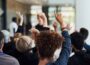 A diverse audience sits facing a speaker at a community event, with several attendees raising their hands to ask questions or participate in the discussion. The speaker is intentionally out of focus, emphasizing audience participation and engagement.