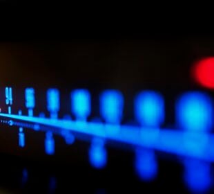 Close-up of an illuminated FM radio tuner dial, with blue frequency markings and a glowing orange indicator positioned near 92 on the scale. The image uses shallow focus, leaving much of the dial softly blurred against a dark background.