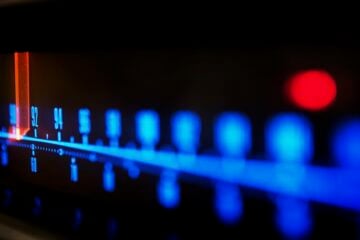 Close-up of an illuminated FM radio tuner dial, with blue frequency markings and a glowing orange indicator positioned near 92 on the scale. The image uses shallow focus, leaving much of the dial softly blurred against a dark background.
