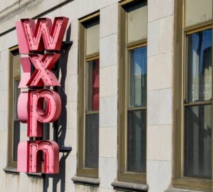 Exterior of a stone office building with a large vertical red neon sign spelling “WXPN” mounted between rows of tall rectangular windows. Sunlight illuminates the façade, casting shadows from the sign onto the building.