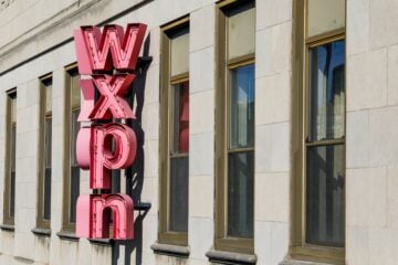 Exterior of a stone office building with a large vertical red neon sign spelling “WXPN” mounted between rows of tall rectangular windows. Sunlight illuminates the façade, casting shadows from the sign onto the building.