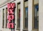 Exterior of a stone office building with a large vertical red neon sign spelling “WXPN” mounted between rows of tall rectangular windows. Sunlight illuminates the façade, casting shadows from the sign onto the building.