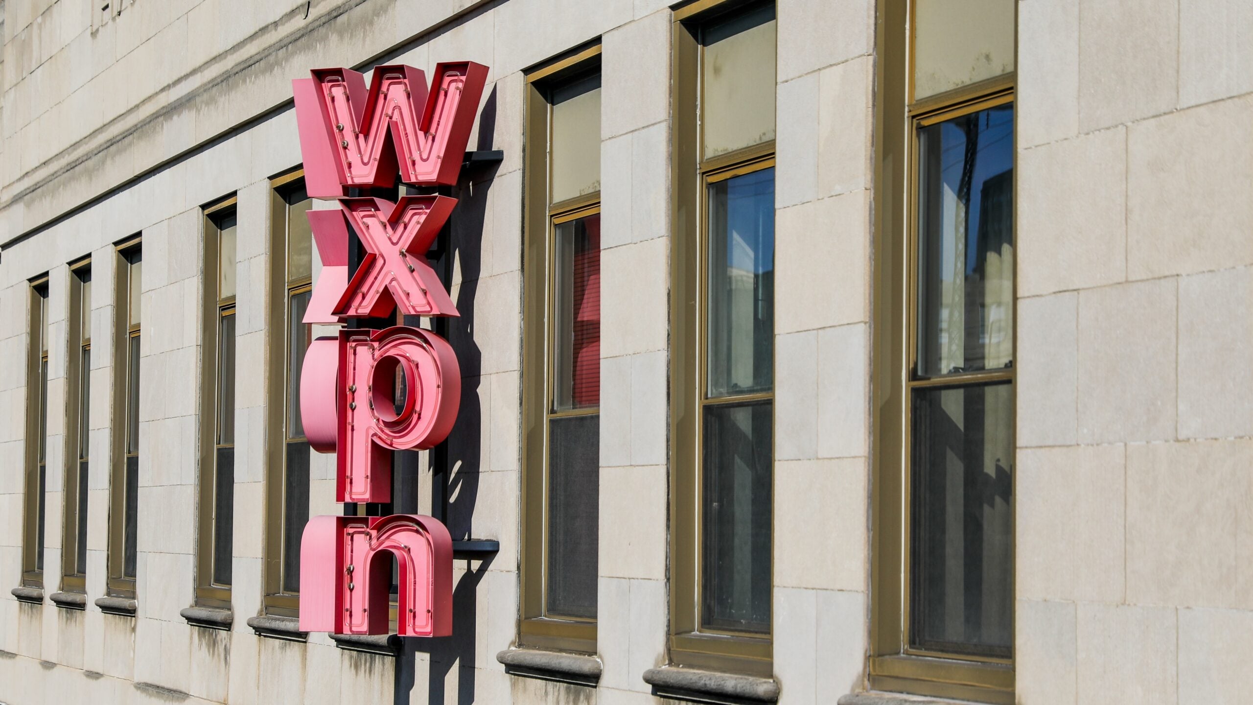 Exterior of a stone office building with a large vertical red neon sign spelling “WXPN” mounted between rows of tall rectangular windows. Sunlight illuminates the façade, casting shadows from the sign onto the building.