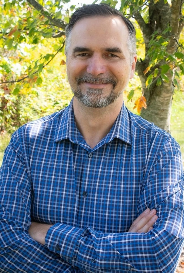 Frank Mueller, station manager of Utah Public Radio, smiles and stands with his arms crossed in front of leafy trees, wearing a blue plaid shirt.
