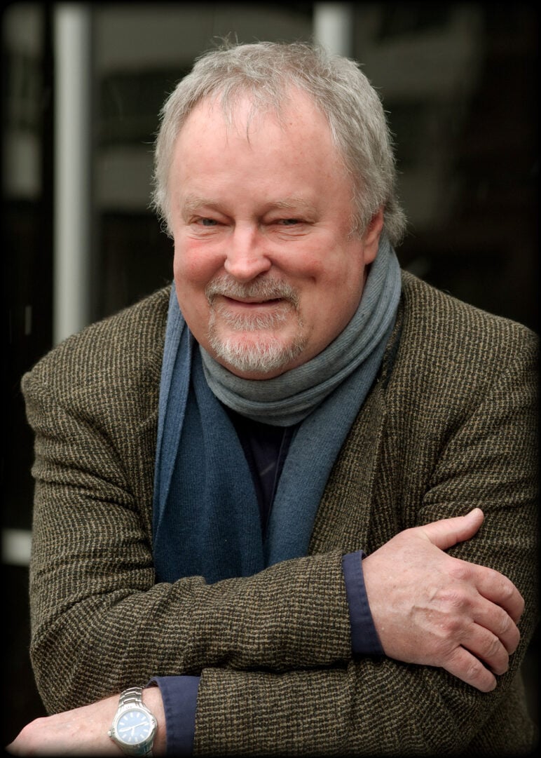 With gray hair and a short beard, Bill McGlaughlin smiles and wears a tweed jacket and layered blue and gray scarves, resting his crossed arms in front of a dark background.