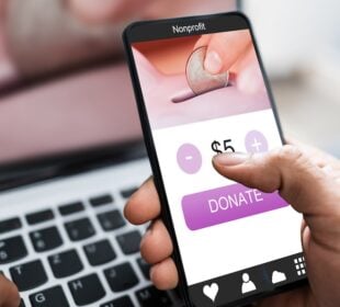Stock image of a hand clicking on a $5 donate button on their phone to a nonprofit with a computer in the background.