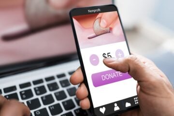 Stock image of a hand clicking on a $5 donate button on their phone to a nonprofit with a computer in the background.