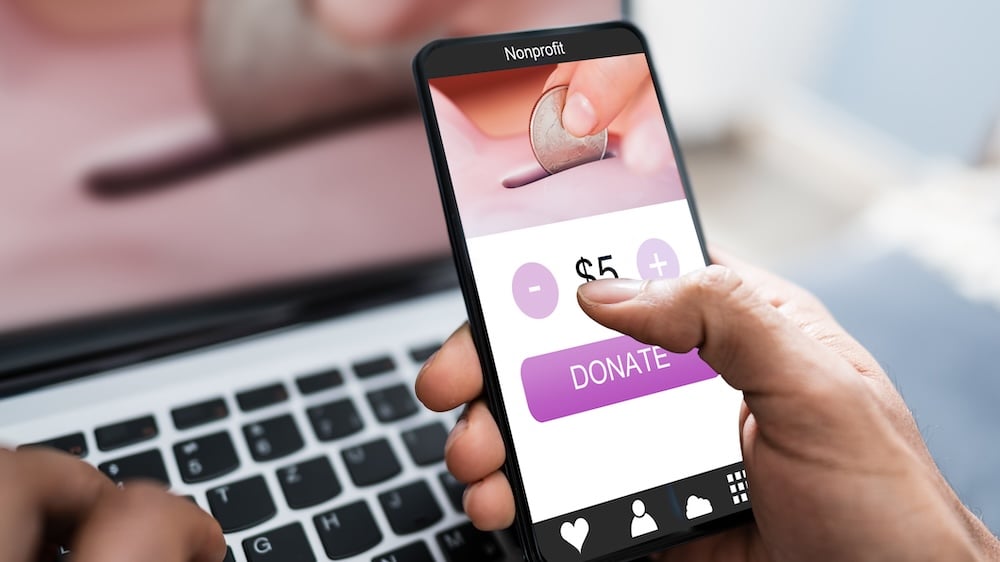 Stock image of a hand clicking on a $5 donate button on their phone to a nonprofit with a computer in the background.