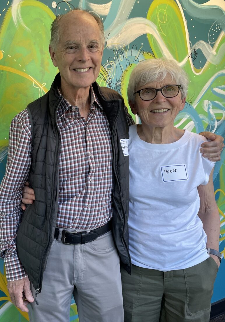 KNKX donors James and Birte Falconer stand smiling with their arms around each other in front of a colorful abstract mural painted in shades of green, blue and white. James wears a checkered button-down shirt and light-colored pants. Birte wears glasses, a white T-shirt and olive-green pants. Both wear handwritten name tags.
