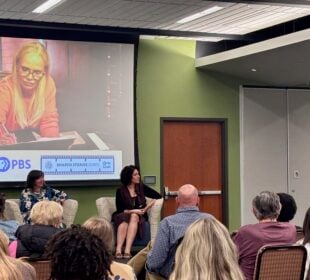 A woman speaks into a microphone at a PBS Utah community event as three panelists sit before an audience. A large screen behind them displays an image from “Finding Your Roots” and logos for PBS Utah and the Children’s Service Society of Utah.