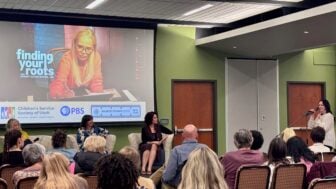 A woman speaks into a microphone at a PBS Utah community event as three panelists sit before an audience. A large screen behind them displays an image from “Finding Your Roots” and logos for PBS Utah and the Children’s Service Society of Utah.