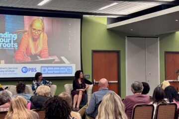 A woman speaks into a microphone at a PBS Utah community event as three panelists sit before an audience. A large screen behind them displays an image from “Finding Your Roots” and logos for PBS Utah and the Children’s Service Society of Utah.
