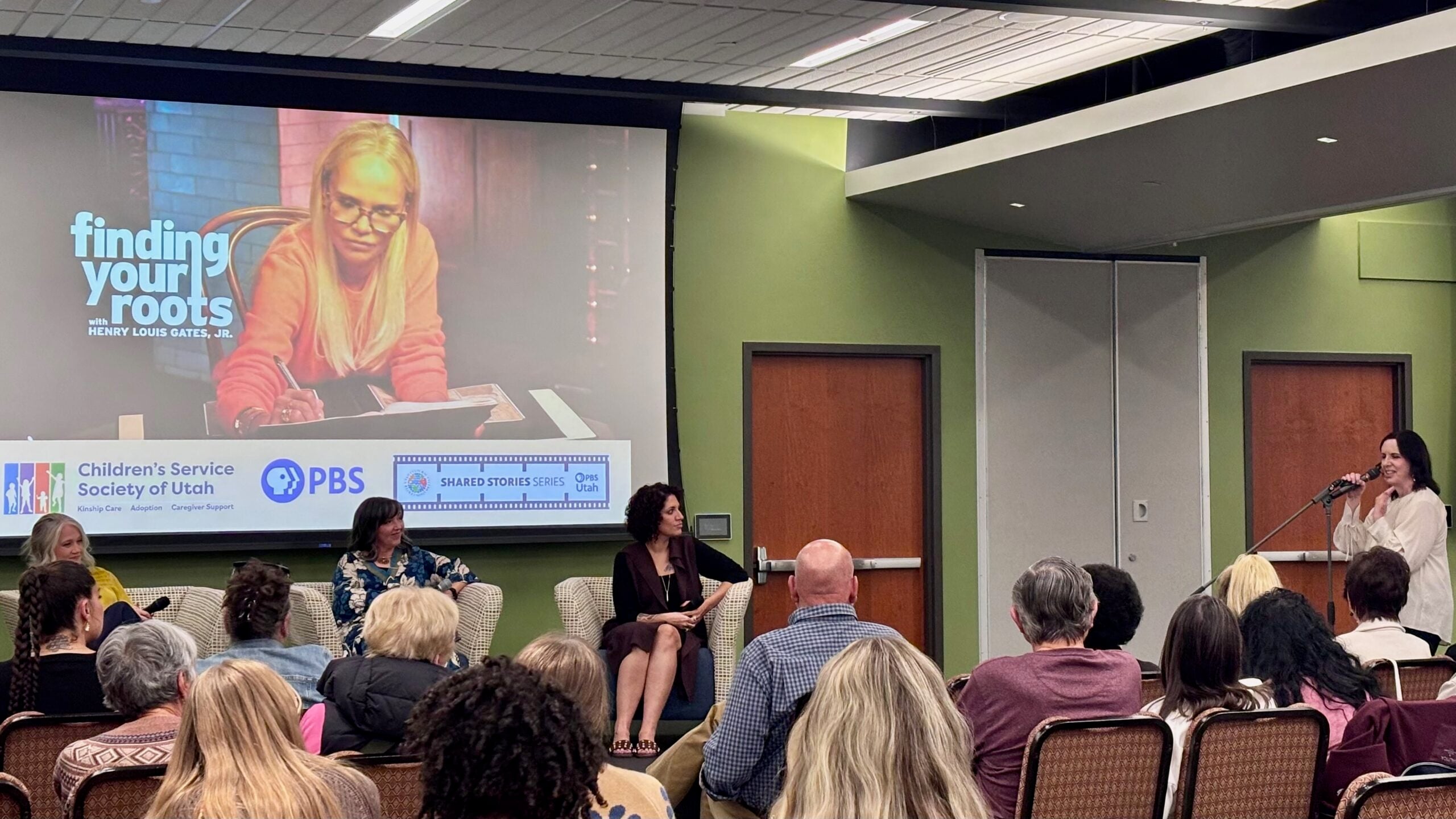 A woman speaks into a microphone at a PBS Utah community event as three panelists sit before an audience. A large screen behind them displays an image from “Finding Your Roots” and logos for PBS Utah and the Children’s Service Society of Utah.