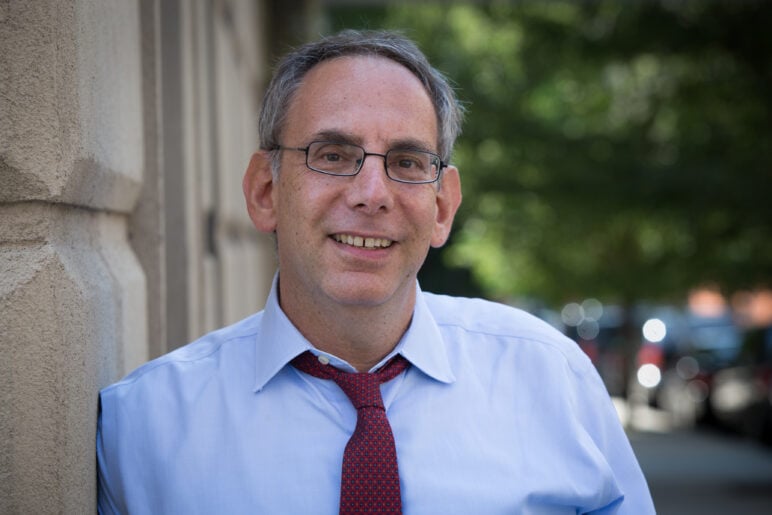 StoryCorps Founder and President Dave Isay smiles while leaning against a stone building, wearing glasses, a light blue shirt and a loosened red tie.