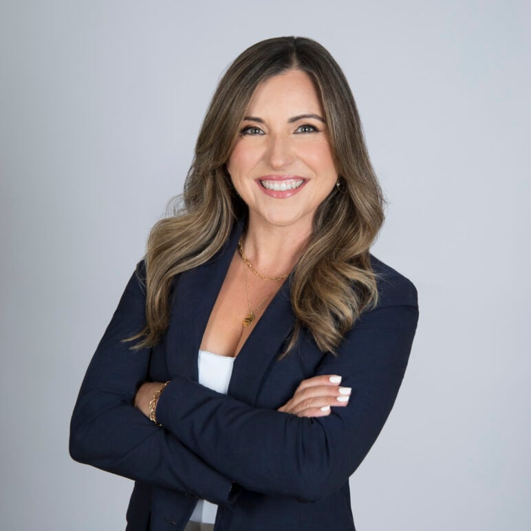 WJCT CEO Karen Feagins smiles and poses with her arms crossed, wearing a navy blazer and gold jewelry against a light gray background.